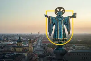 Blick auf das Münchner Kindl mit einem gelben Wappenrahmen über der Skyline Münchens