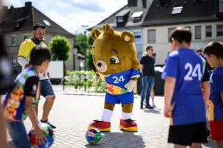 EM-Maskottchen Albärt umgeben von Kindern auf einem Schulhof bei Sonnenschein