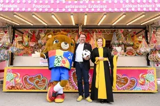 European Championship mascot Albärt, sports officer Florian Kraus and Münchner Kindl Franziska Inselkammer in front of a stand at the spring festival